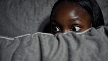Surprised person peeking over cozy blanket with wide eyes in dimly lit room