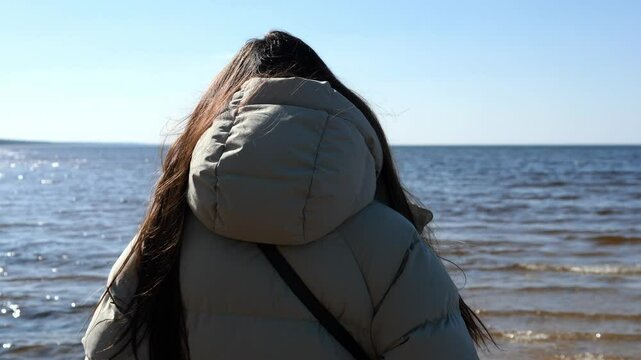Young woman in warm puffer jacket stands on sandy beach, watching gentle waves roll across calm sea. Sandy beach stretches under clear sky, as footsteps mark soft surface of sandy beach. 