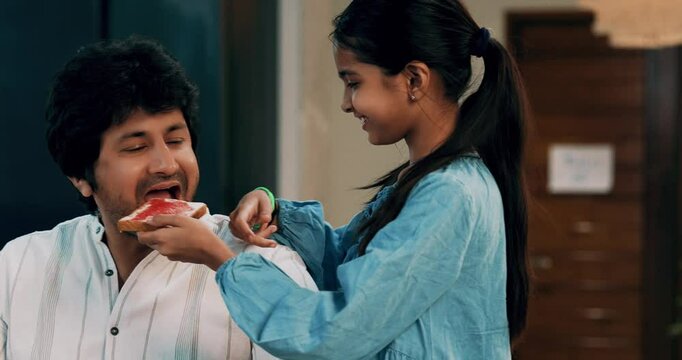 Indian Asian young father and his little daughter sitting together at the dining table in a stylish modern home, girl sharing bread jam with dad, bonding over a nutritious morning meal