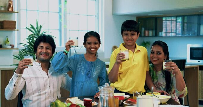 Indian Asian young parents and two kids, sitting together at the breakfast table in modern home, happily holding glasses of A2 quality milk and showing them to the camera to promote a healthy diet