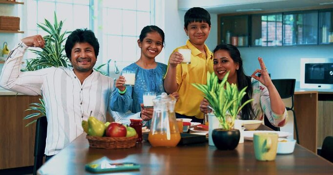 Indian Asian young parents and two kids, sitting together at the breakfast table in modern home, happily holding glasses of A2 quality milk and showing them to the camera to promote a healthy diet