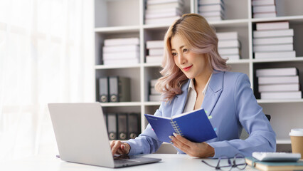Professional businesswoman reading notebook while using laptop mouse at modern office desk with bookshelf background © Pichsakul