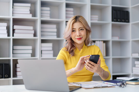 Businesswoman using smartphone at office desk with laptop, coffee cup and sticky notes on window..