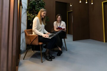 Fototapeta premium Two women collaborate in a modern workspace while using a laptop and a book in an indoor setting during daylight hours