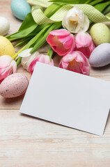 Easter greeting card lying near colorful eggs and pink tulips on white wooden table