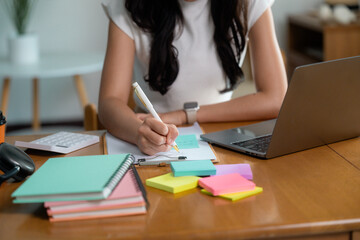 Asian businesswoman writing notes while working from home office using laptop and colorful sticky...