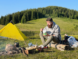 Man tourist eating from food pouch with green spoon, sits on log near campfire. Yellow tent on...