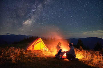 Couple having rest by campfire near their illuminated tent beneath stunning star-filled sky. Milky...