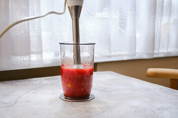 Blending fresh tomatoes in a clear glass container using an immersion blender in the kitchen