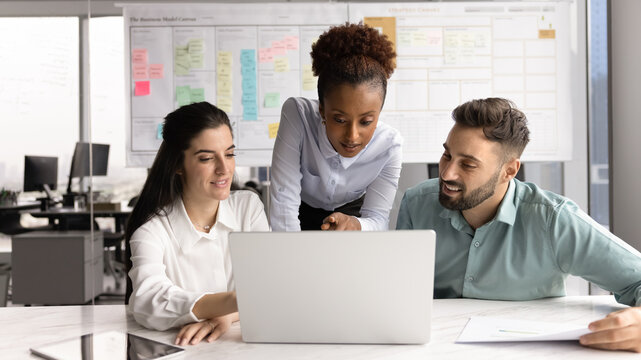 Young African business leader woman and two Caucasian managers discussing Internet marketing, sales growth at laptop, meeting at computer for teamwork, brainstorming, talking at pc gadget in office