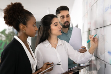 Serious Latin business leader woman writing tasks on Kanban white board, talking to young diverse...