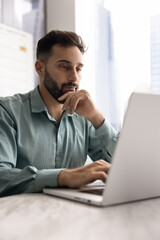 Focused pensive handsome Latin businessman working at laptop in office, touching face and chin in...