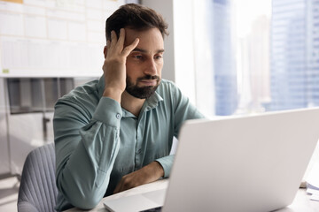 Tired annoyed Latin businessman touching head at laptop, reading or watching about problem,...