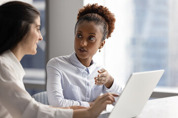 Serious young African businesswoman talking to positive female colleague at laptop. Diverse...