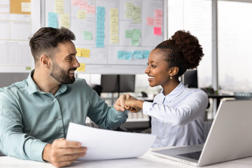 Happy diverse project managers celebrating business success and giving fist bump, having fun at workplace table, counting income from successful project, holding financial growth paper report