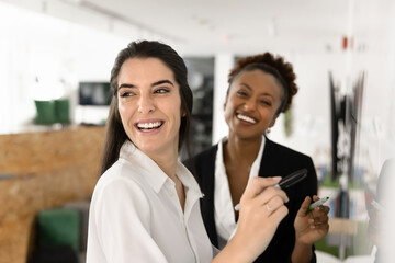 Obraz premium Cheerful diverse female coworkers writing teamwork plan on whiteboard, looking at camera and away, smiling, laughing. Office friends brainstorming, enjoying working together