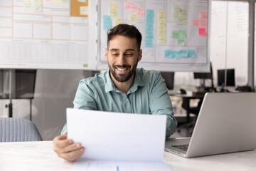Positive satisfied Hispanic business professional man in casual reading paper report at workplace with laptop, checking legal financial documents, letter with good news, smiling