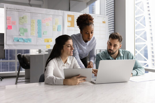 Diverse business team of young colleagues discussing online startup project at laptop and tablet, meeting for teamwork, analyzing electronic reports in co-working scrum management space
