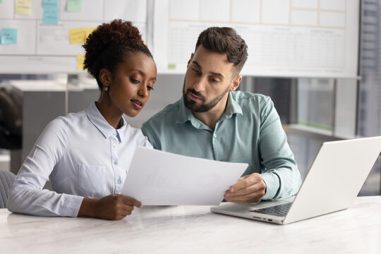 Two serious focused multiethnic male and female coworkers reviewing financial legal document, discussing sales marketing reports, brainstorming on project, doing paperwork at office table