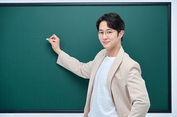 A young male teacher in a suit is holding a piece of chalk and giving a lecture in front of a blackboard