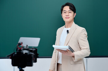 A young male instructor in a suit is holding a book and delivering an online lecture in front of...