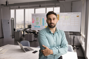 Fototapeta premium Confident handsome Hispanic executive man posing in office space alone, keeping arms folded, looking at camera. Corporate business portrait of successful company leader, entrepreneur