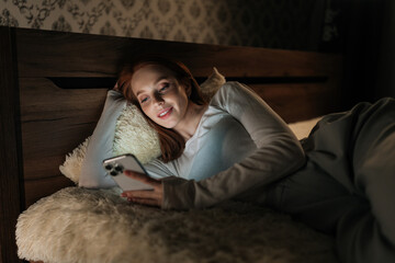 Attractive young woman lying in bed at night, using mobile phone smiling looking to screen,...