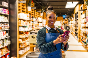Saleswoman using smartphone in organic supermarket with shelves full of products