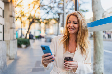 Woman Smiling While Waiting at Bus Stop in City - Happy woman with coffee and phone waiting at a public transport stop in an urban setting.