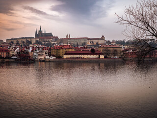 Naklejka premium Panoramic view of Prague Castle and historic buildings along the Vltava River at dusk, with calm water and moody sky.