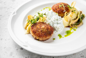 A delicious plate featuring golden-brown fish patties accompanied by creamy tzatziki, mashed potatoes, and garnished with fresh herbs and crispy vegetables.