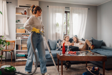 Mother vacuuming living room while daughters relax on sofa