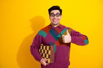 Joyful young man with glasses in colorful sweater holding a chessboard and showing a thumbs-up...