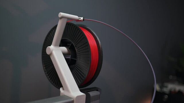 A roll of red high quality 3D printing filament mounted on a modern 3D printer device, closeup. Consumer grade thermoplastic materials used for object product prototyping and design