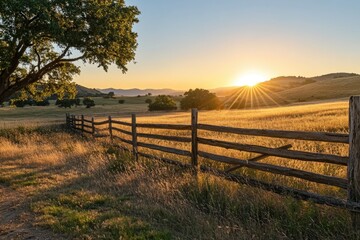 The sun is setting over a rural landscape with grain fields, hills in the distance, and a wooden fence leading towards it.