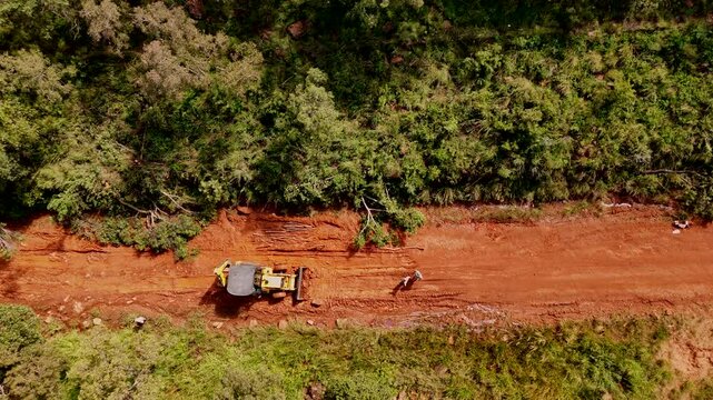 A construction worker operates a TLB (tractor-loader-backhoe) to fix a muddy road in a forested area, with a dirt path and trees around