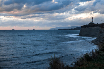 Gelendzhik resort. A lighthouse is visible on the shoreline of a body of water. The sky is cloudy and the water is calm