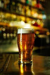 Cold beer in pint glass with condensation placed on shiny bar surface in front of bottle shelf. Concept of alcohol, festival, beer, culture, pub, bar, celebration, Friday mood.