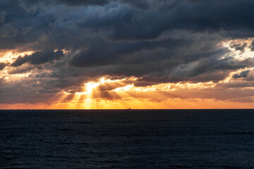 The sky is dark and cloudy with a few rays of sunlight peeking through. The ocean is calm and the horizon is visible