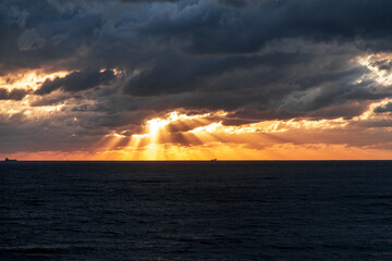 The sky is dark and cloudy with a few rays of sunlight peeking through. The ocean is calm and the horizon is visible