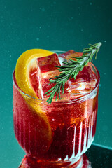 Negroni-style cocktail with rosemary and grapefruit on teal background. Bold cocktail with ruby red ice cubes, rosemary sprig and grapefruit slice. Concept of nightlife, party, bar culture, alcohol.