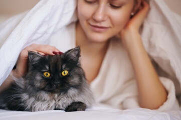 Woman lying beside fluffy cat under blanket cherishing intimate calm, sharing affectionate gaze,...
