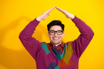Smiling young man with retro-style sweater forming a house roof gesture with his hands against a yellow background.