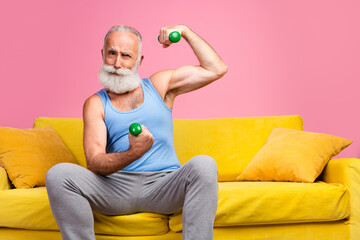 Senior man with a white beard flexing with weights on a yellow sofa against a pink background