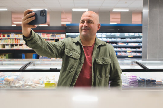 Smiling mature man taking a selfie with his smartphone while shopping for frozen food in a supermarket, capturing a moment of his grocery shopping experience