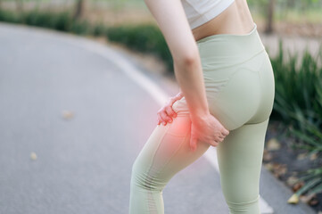 young Asian woman experiencing pain her thigh during exercise outdoors, holding her leg with discomfort. She is wearing light green leggings and white top, standing paved path surrounded by