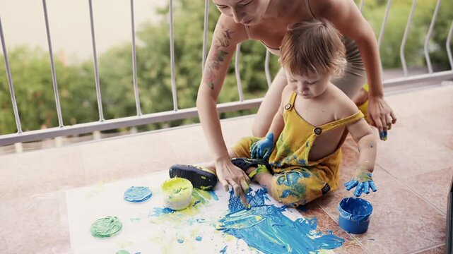 Young mother with her little boy mixing colors at hands yo leave beautiful handprints on the paper. Mother and child concept. Slowmotion. Mom amd child spend time together. Family, parenthood concept