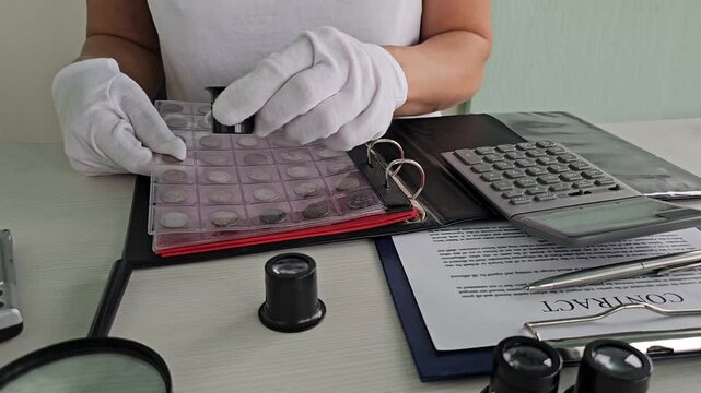 Coin collector examines rare coins with magnifying glass and tools in organized display at a desk