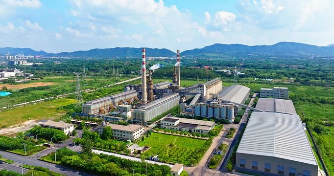 Aerial view of an industrial plant with chimneys releasing steam, surrounded by green fields, trees, and mountains.