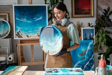 Happy female artist artisan in protective equipment works on circular resin epoxy piece with ocean waves in workshop. Creative craftswoman, blending colors, passion and hobby at home, do it yourself